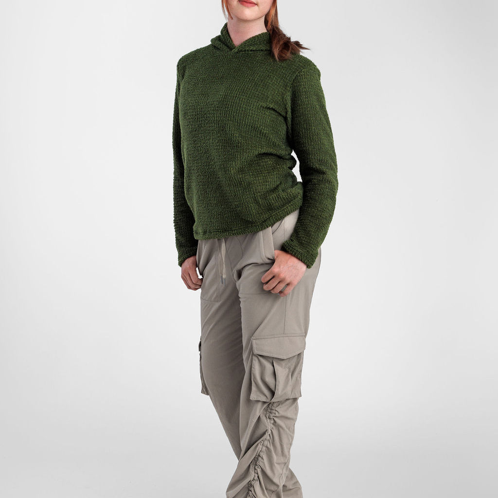 A woman modeling a dark green Alpha 120 Hoodie and light beige cargo pants with ruffled details, standing against a light gray studio background.