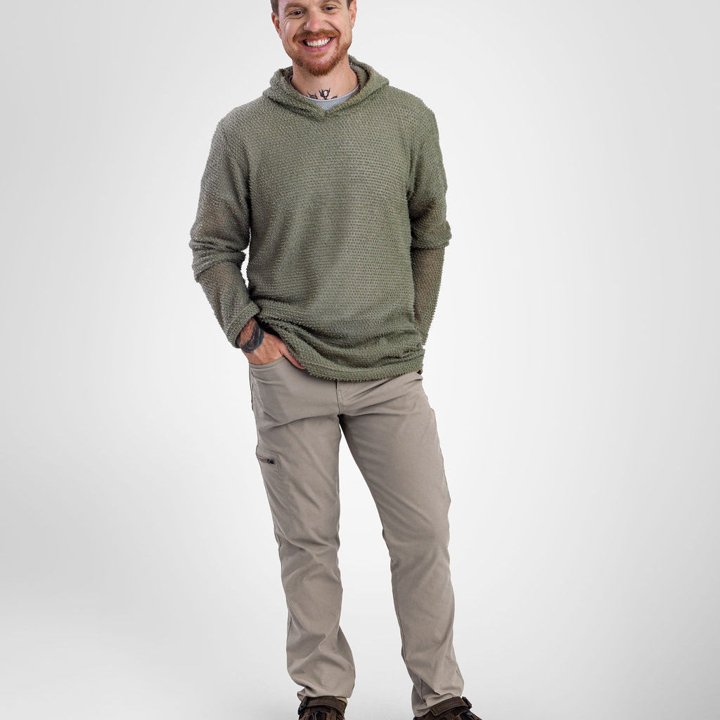 A man wearing an olive green Alpha 60 hoodie hoodie, beige cargo pants, and brown hiking shoes, standing against a light gray studio background.