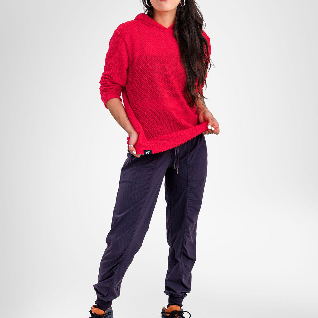 A woman modeling a bright red long-sleeve hoodie and dark navy jogger pants with black hiking boots, standing against a light gray studio background.