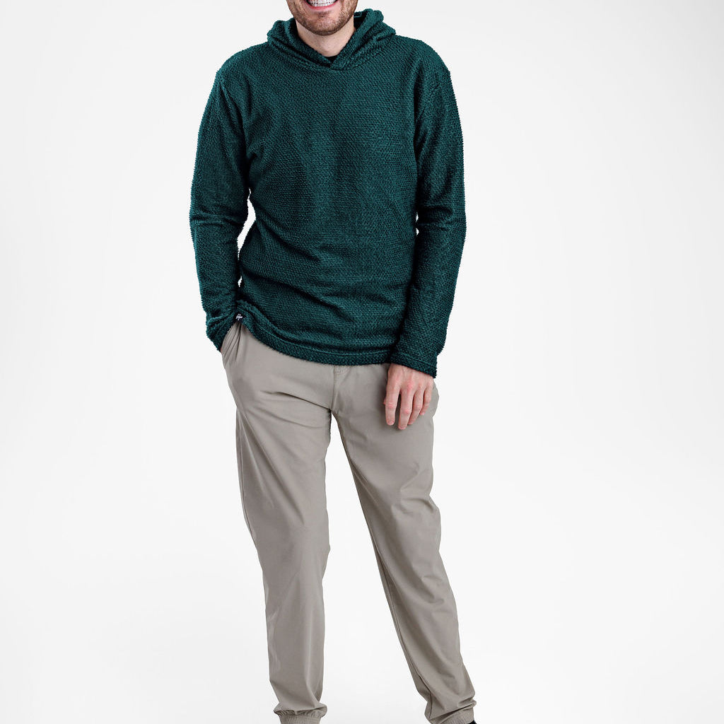 A man wearing a dark green textured Alpha 90 hoodie, light gray pants, and black sneakers, standing against a plain light gray studio background.