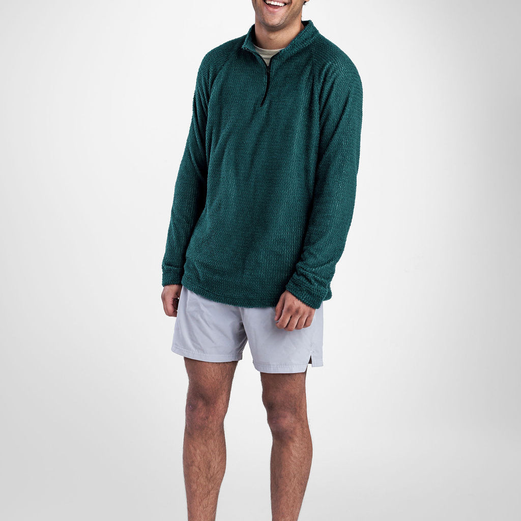 A man wearing a dark green quarter-zip Polartec Alpha Direct Hoodie, white athletic shorts, and gray running shoes, standing against a light gray studio background.