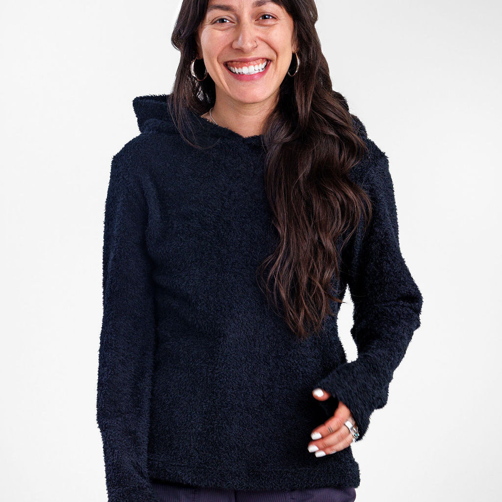A smiling woman with long dark hair modeling a black Alpha 90 hoodie and wearing dark purple pants, standing against a plain white studio background.