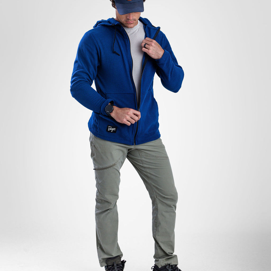 A man wearing a royal blue zip-up highlander hoodie, gray pants, black shoes, and a navy cap with an orange logo, standing against a light gray studio background.