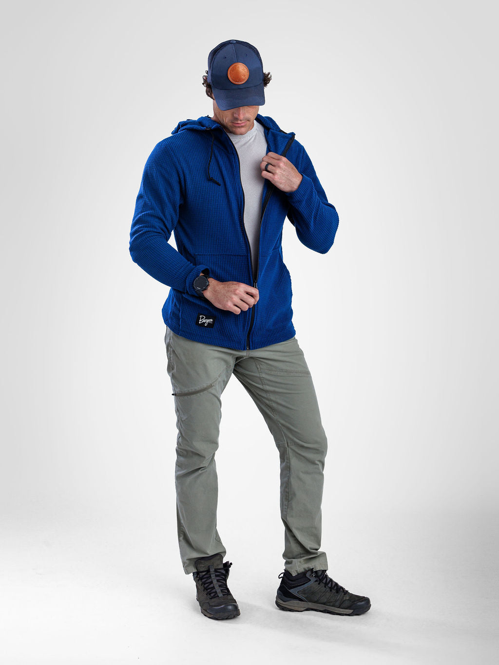 A man wearing a royal blue zip-up hooded jacket, gray pants, black shoes, and a navy cap with an orange logo, standing against a light gray studio background.