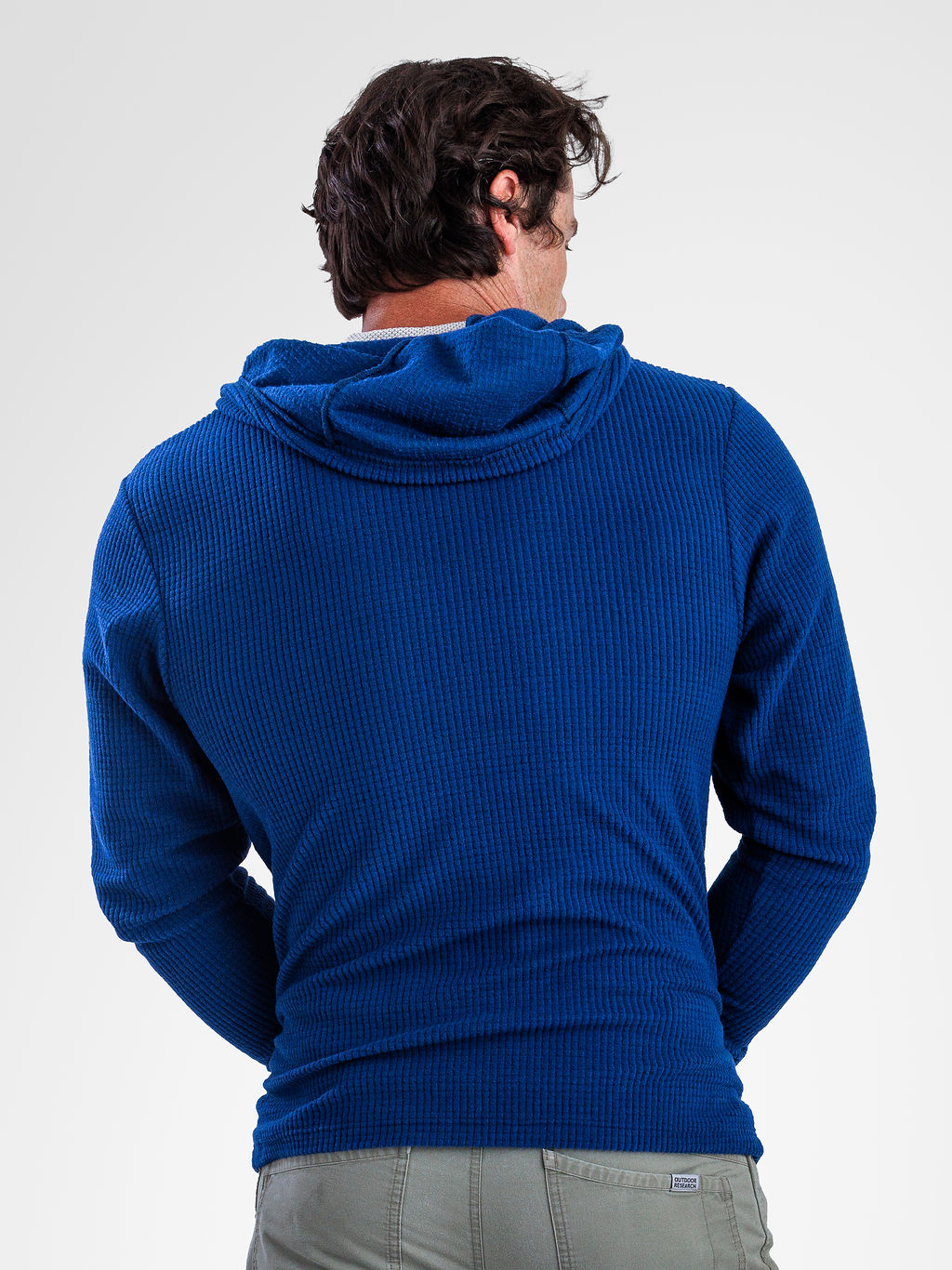 A man viewed from behind wearing a royal blue ribbed hooded sweater and light gray pants against a plain light gray studio background.