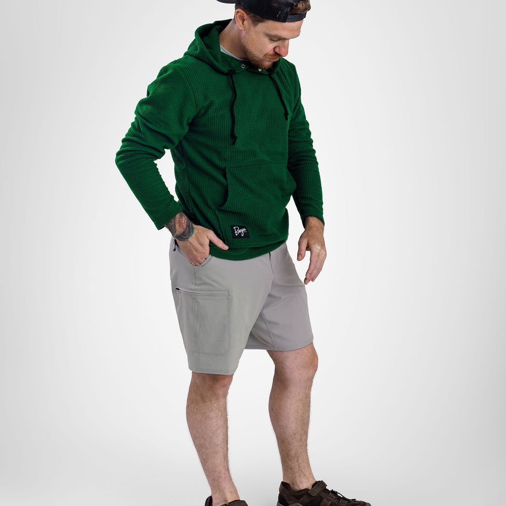 A man wearing a dark green Highlander fleece hoodie, light gray shorts, and brown hiking shoes, standing against a light gray studio background.
