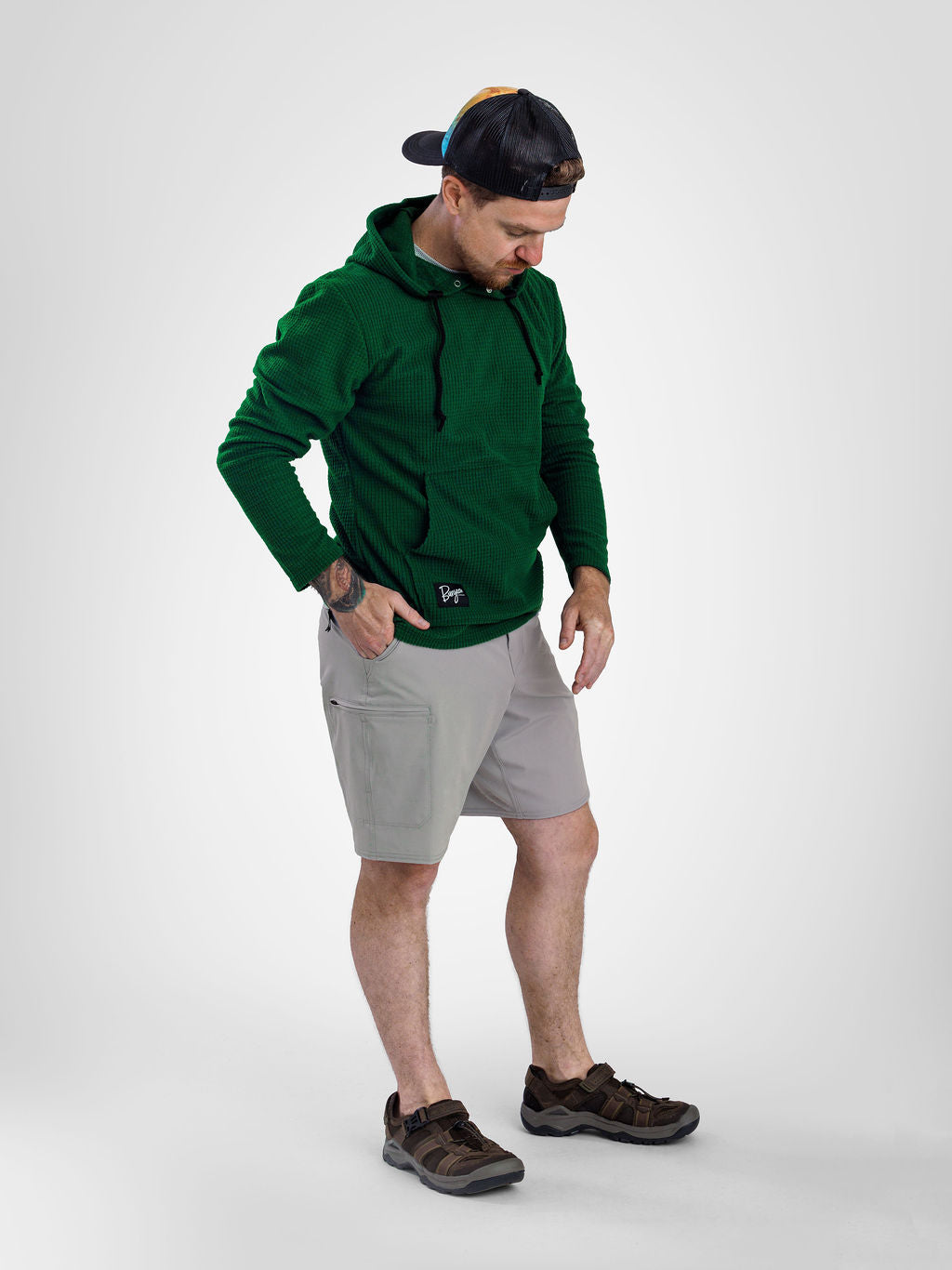 A man wearing a dark green Highlander fleece hoodie, light gray shorts, and brown hiking shoes, standing against a light gray studio background.