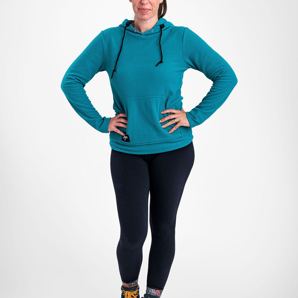 A woman modeling a teal Highlander hoodie with black drawstrings, paired with black leggings and hiking boots, standing against a light gray studio background.