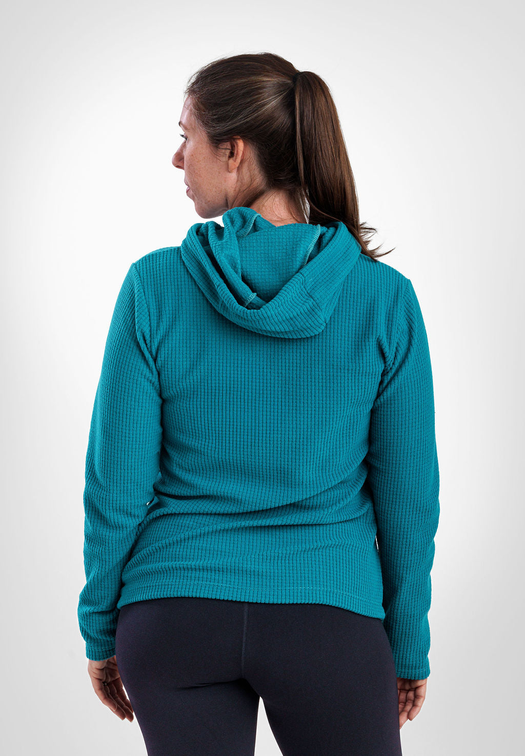 A woman shown from the back wearing a teal Highlander Hoodie and dark leggings against a light gray studio background.