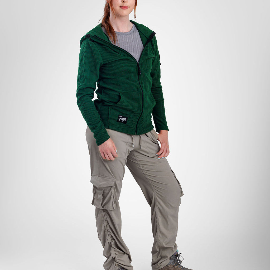 A woman modeling a dark green zip-up fleece jacket, paired with light gray cargo pants and hiking boots, against a light gray studio background.