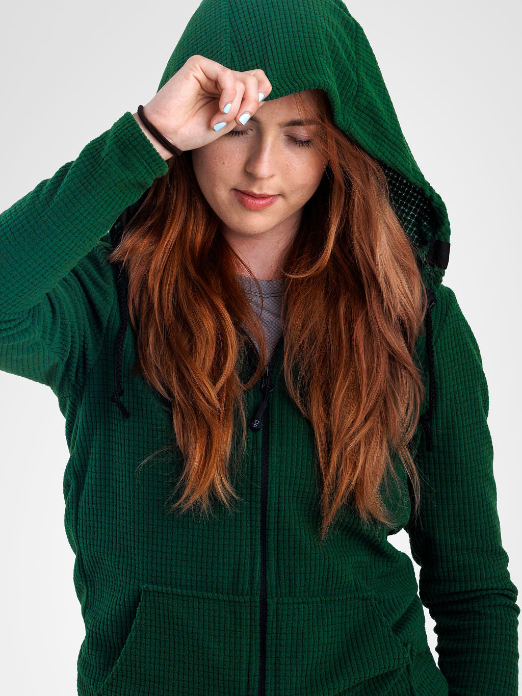 A woman wearing a dark green waffle-knit zip-up hoodie with the hood up, shown against a plain light gray studio background.