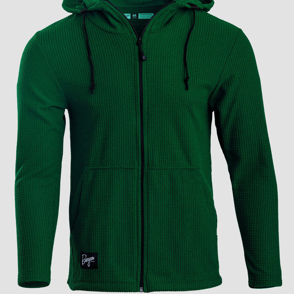 A dark green waffle-knit zip-up hoodie with a drawstring hood and a small black brand patch on the lower left hem, shown against a light gray background.