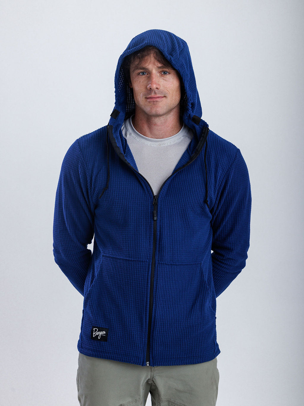 A man wearing a royal blue ribbed zip-up hoodie with a black zipper and a small black brand patch on the lower left hem, paired with a light gray t-shirt and khaki pants, against a plain light gray background.