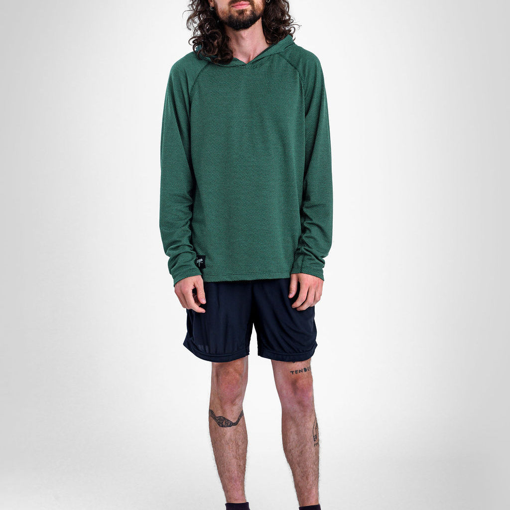 A man with long curly hair and a beard stands against a light gray studio background wearing a dark green long-sleeve Sunseeker Hoodie, dark navy athletic shorts, dark socks, and gray running shoes.