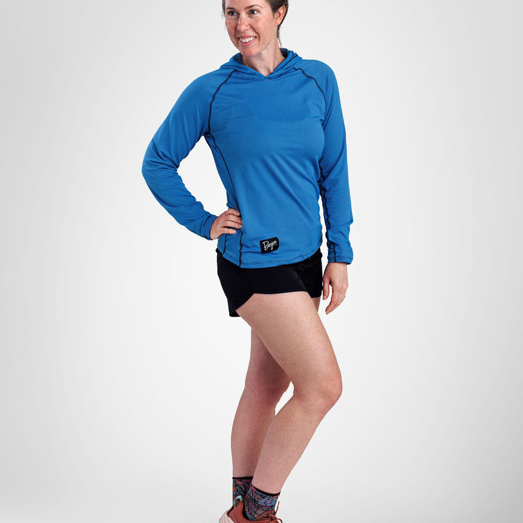 A woman modeling a bright blue long-sleeve sun hoodie with black running shorts, patterned socks, and brown trail running shoes against a light gray studio background.