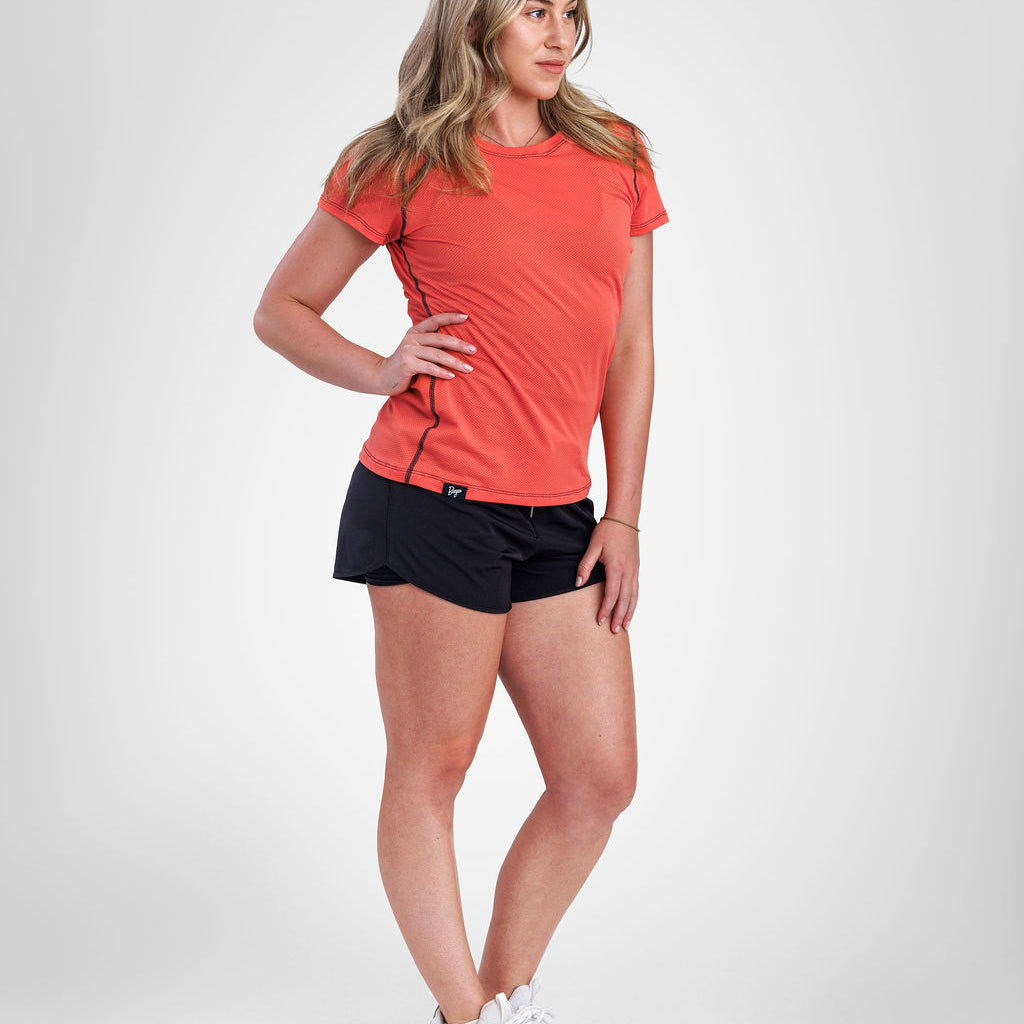 A woman modeling a coral orange short-sleeve athletic t-shirt and black athletic shorts, paired with white sneakers, against a light gray studio background.