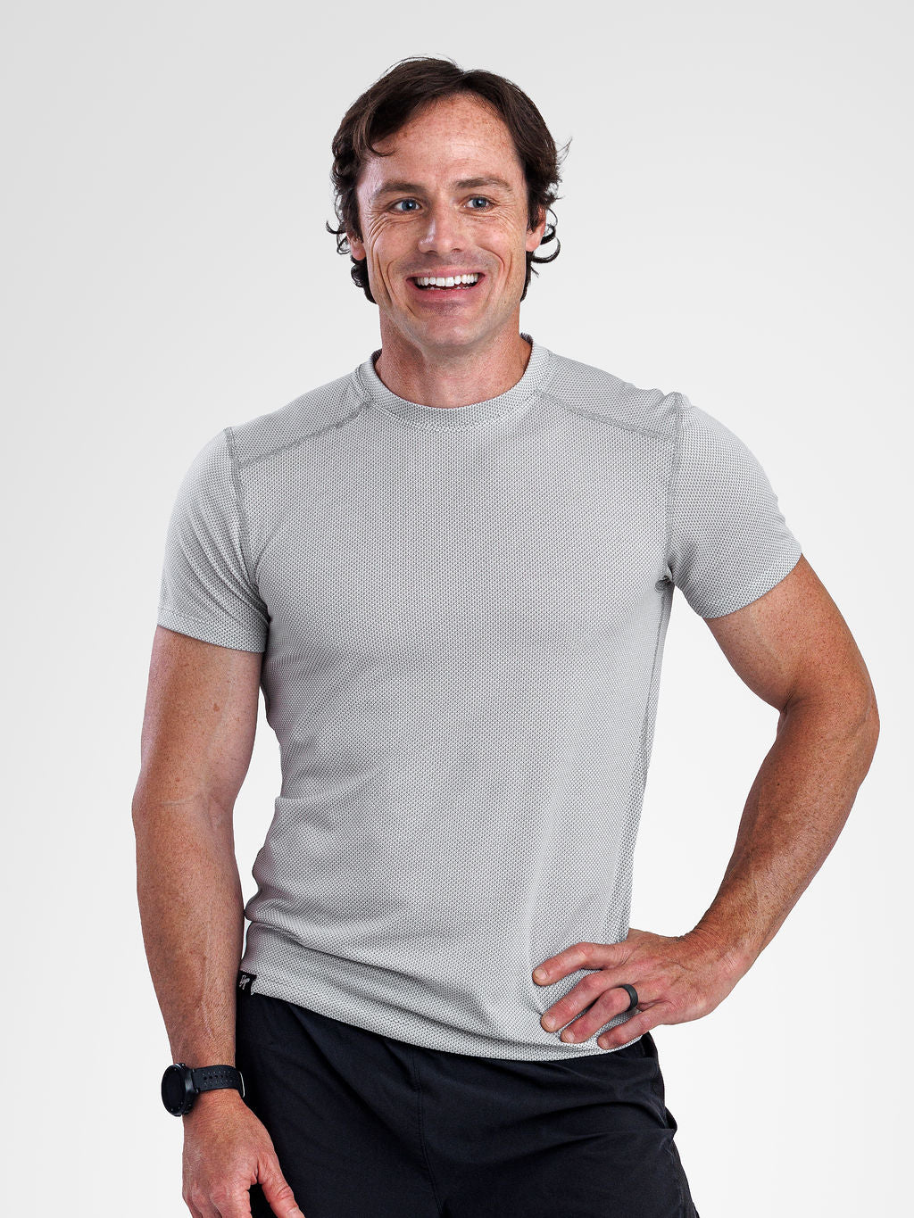 A smiling man wearing a light gray short-sleeve athletic t-shirt and black pants, standing against a light gray studio background.