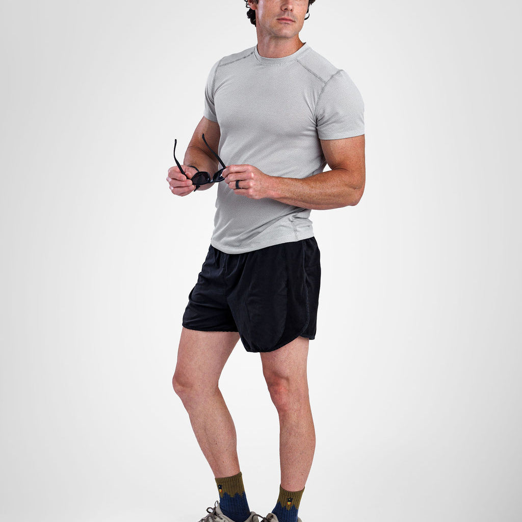 A man modeling a light gray short-sleeve athletic t-shirt, black athletic shorts, a navy blue baseball cap with an orange logo, olive green socks, and gray trail running shoes against a light gray studio background.