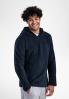 A smiling man with curly dark hair wearing a dark navy Alpha 120 hoodie and light gray pants, standing against a plain light gray studio background.