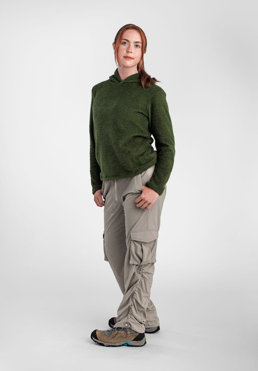 A woman modeling a dark green Alpha 120 Hoodie and light beige cargo pants with ruffled details, standing against a light gray studio background.