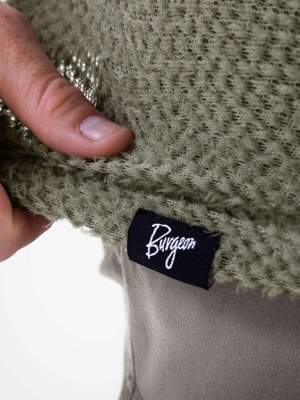 Close-up of a person's hand holding an Alpha 60 hoodie to show the texture and a black fabric brand tag with the name Burgeon in white script.
