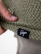 Close-up of a person's hand holding an Alpha 60 hoodie to show the texture and a black fabric brand tag with the name Burgeon in white script.