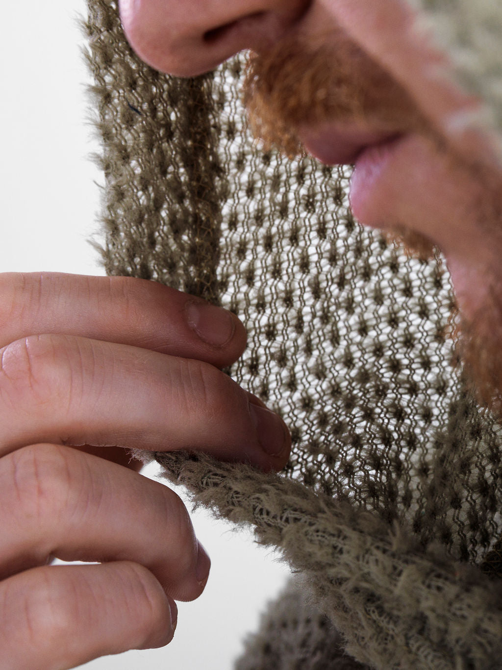 Close-up of a person wearing an Alpha 60 hoodie, with the hand holding the edge of the textured fabric to show its thickness and the nose and lips of the wearer visible behind it.