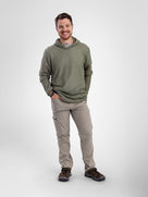 A man wearing an olive green Alpha 60 hoodie hoodie, beige cargo pants, and brown hiking shoes, standing against a light gray studio background.
