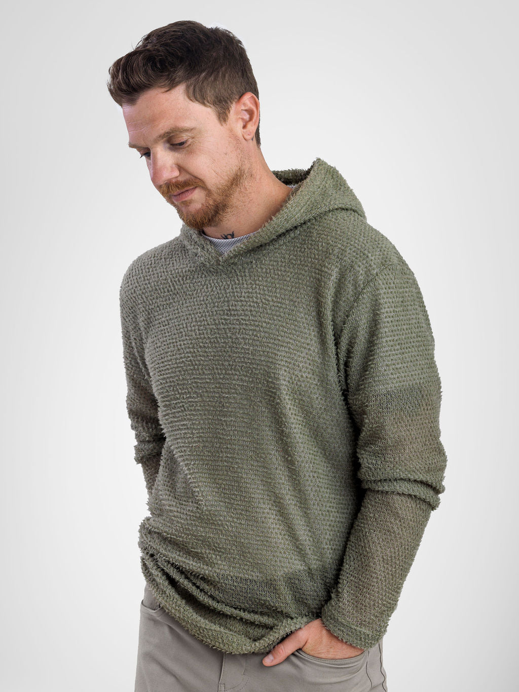 A man wearing a textured olive green Alpha 60 hoodie, looking down and to the side, paired with gray pants, against a plain light gray background.