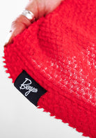 A close-up of a bright red textured Alpha knit fabric with a black fabric label sewn onto the edge, featuring the brand name 'Burgeon' in white script. A hand with a silver ring is holding the fabric.