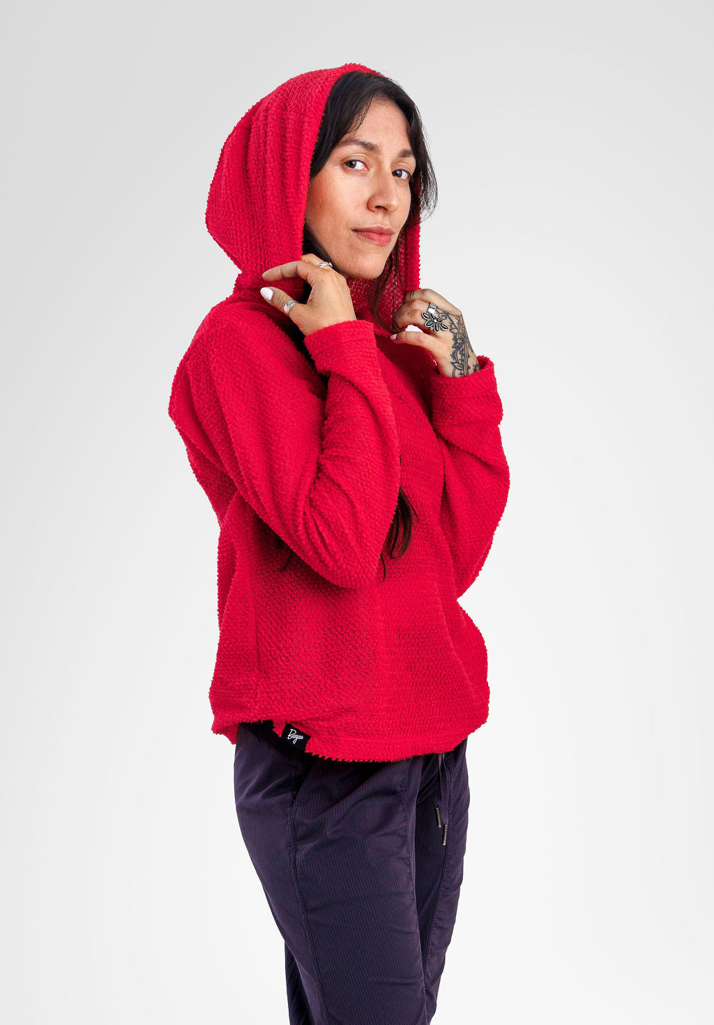 A woman wearing a bright red Alpha fleece hooded sweatshirt and dark navy pants, posing against a light gray studio background.