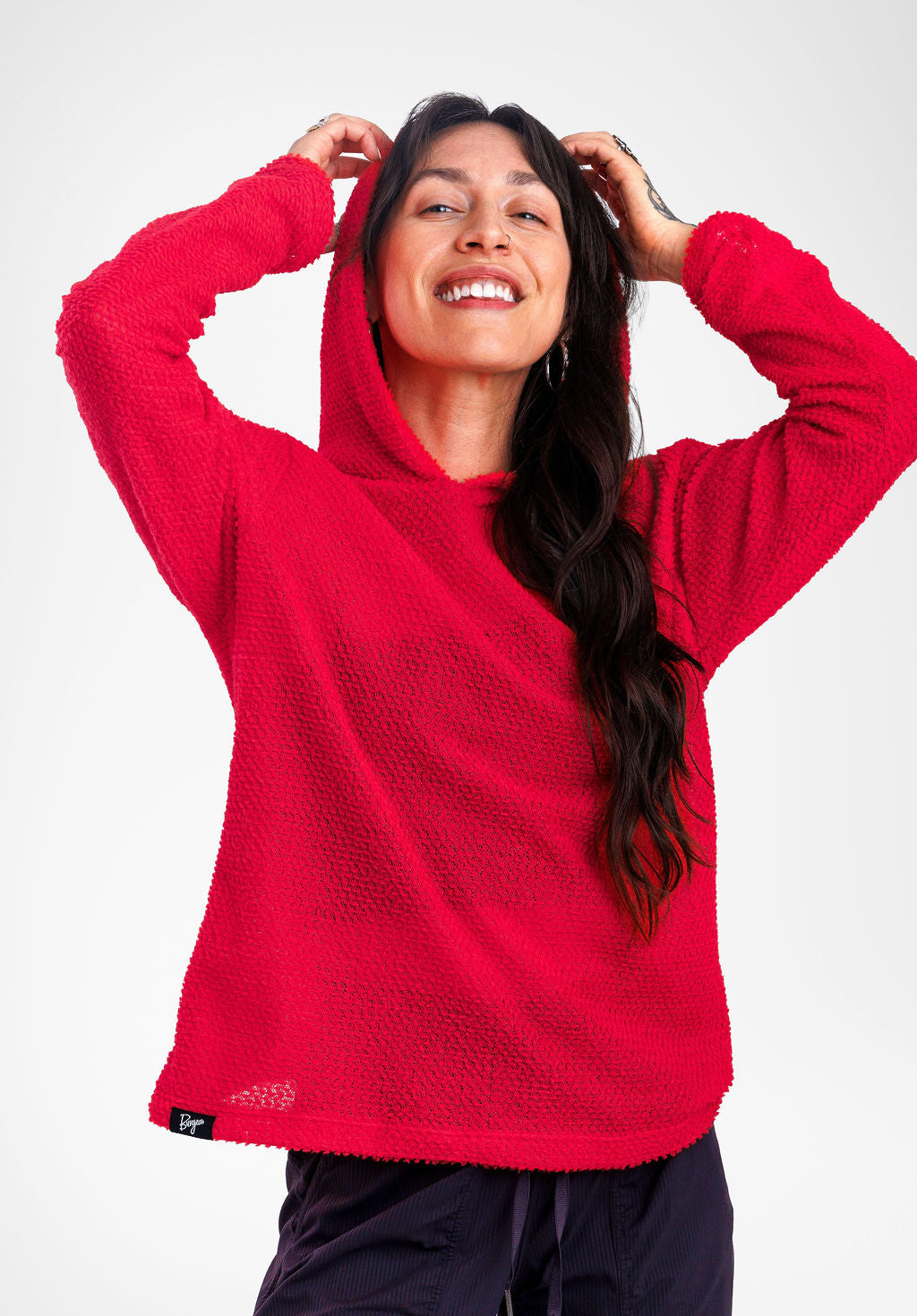 A woman wearing a bright red textured Alpha fleece hoodie with the hood up, posing against a light gray studio background.