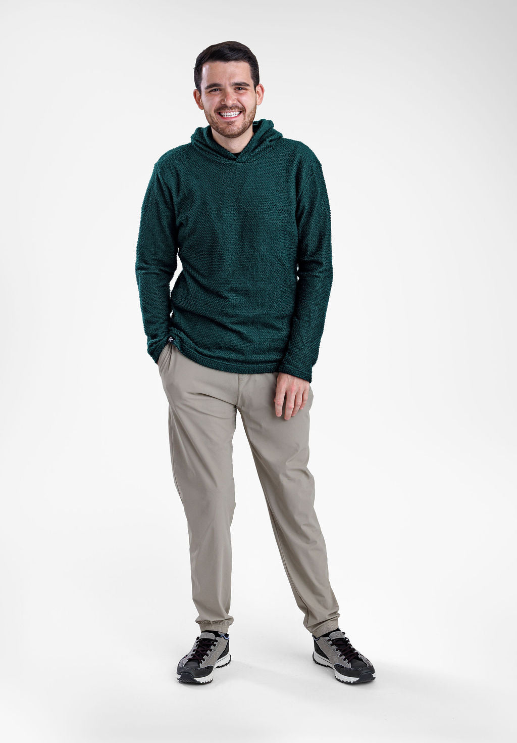 A man wearing a dark green textured Alpha 90 hoodie, light gray pants, and black sneakers, standing against a plain light gray studio background.