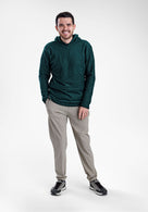 A man wearing a dark green textured Alpha 90 hoodie, light gray pants, and black sneakers, standing against a plain light gray studio background.