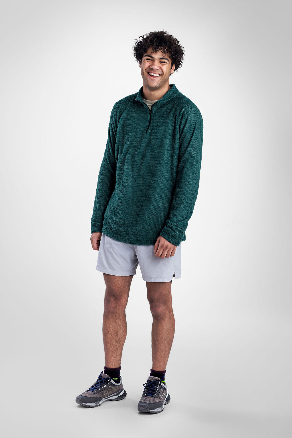 A man wearing a dark green quarter-zip Polartec Alpha Direct Hoodie, white athletic shorts, and gray running shoes, standing against a light gray studio background.