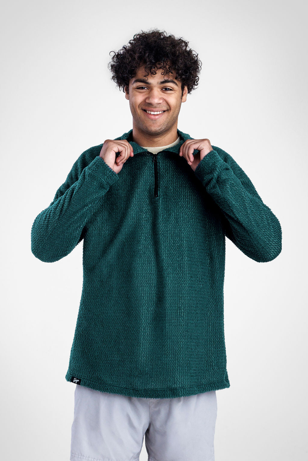A smiling man with curly dark hair wearing a dark green Polartec Alpha Direct Hoodie and light gray pants, posing against a light gray studio background.