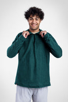 A smiling man with curly dark hair wearing a dark green Polartec Alpha Direct Hoodie and light gray pants, posing against a light gray studio background.