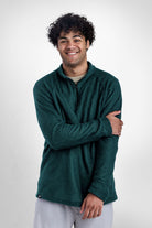 A smiling man with curly dark hair wearing a dark green Polartec Alpha Direct Hoodie and light gray pants, standing against a plain light gray studio background.
