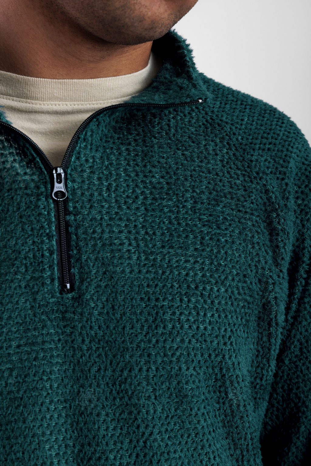 Close-up of a person wearing a dark teal quarter-zip Polartec Alpha Direct Hoodie over a light beige t-shirt, against a plain light gray background.