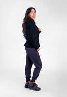 A woman modeling a black Alpha 90 hoodie wearing dark navy jogger pants and hiking shoes, standing against a light gray studio background.
