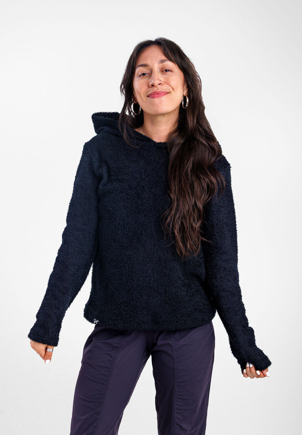 A woman modeling a black Alpha 90 hoodie, wearing dark purple pants against a plain light gray studio background.