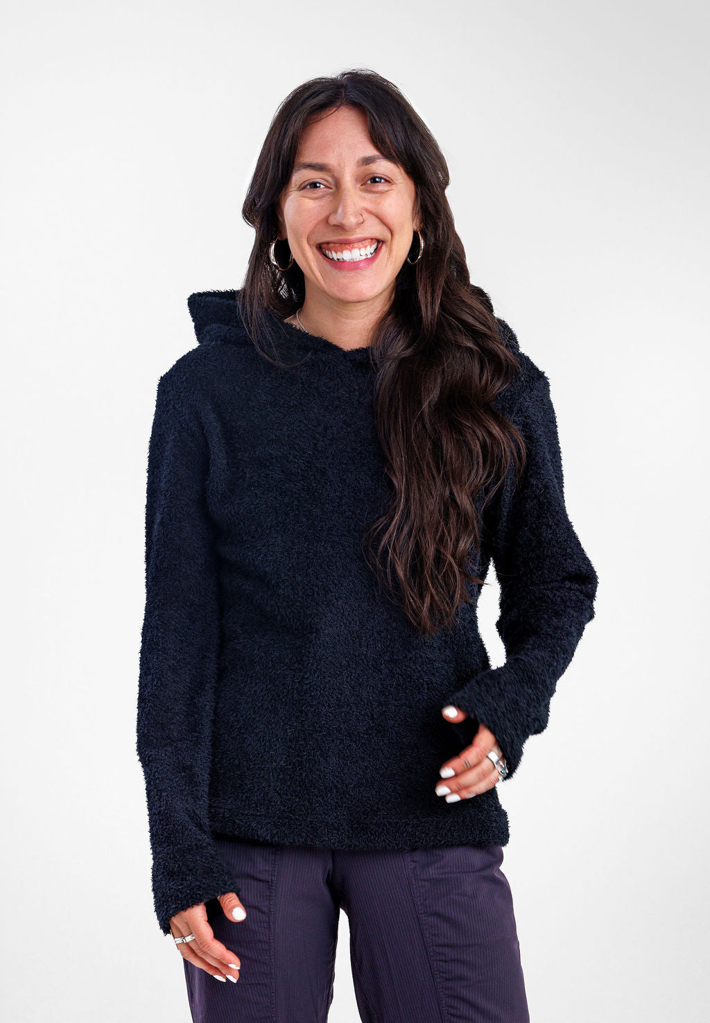 A smiling woman with long dark hair modeling a black Alpha 90 hoodie and wearing dark purple pants, standing against a plain white studio background.