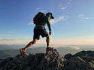 A hiker wearing a green Highlander hoodie, black shorts, and a black backpack struggles to climb a rocky peak under a bright blue sky with distant mountain ranges.
