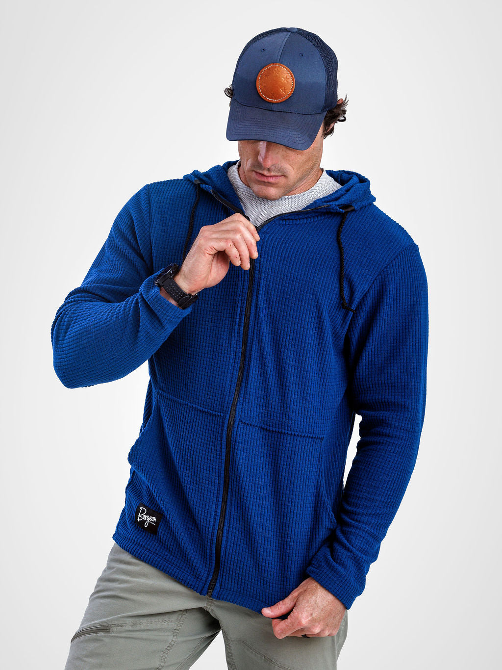 A man wearing a royal blue waffle-knit zip-up hoodie with a drawstring hood, paired with gray pants and a navy blue baseball cap with a circular orange patch, photographed against a light gray studio background.