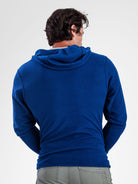 A man viewed from behind wearing a royal blue ribbed hooded sweater and light gray pants against a plain light gray studio background.