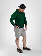 A man wearing a dark green Highlander fleece hoodie, light gray shorts, and brown hiking shoes, standing against a light gray studio background.