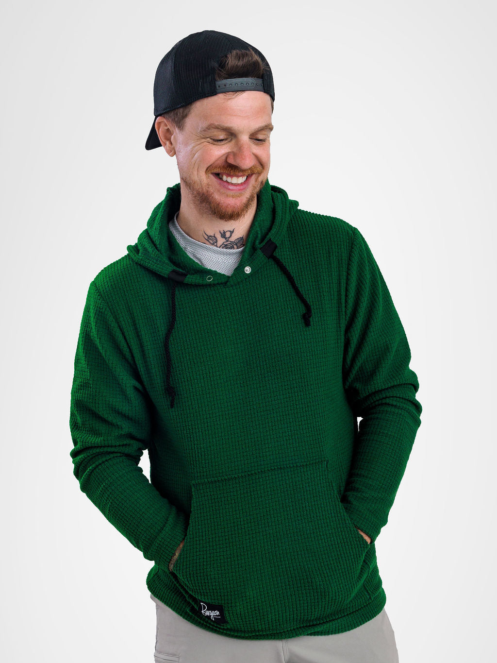 A man wearing a dark green Highlander fleece hoodie with black drawstrings, a black baseball cap backwards, and a gray t-shirt underneath, standing against a light gray studio background.