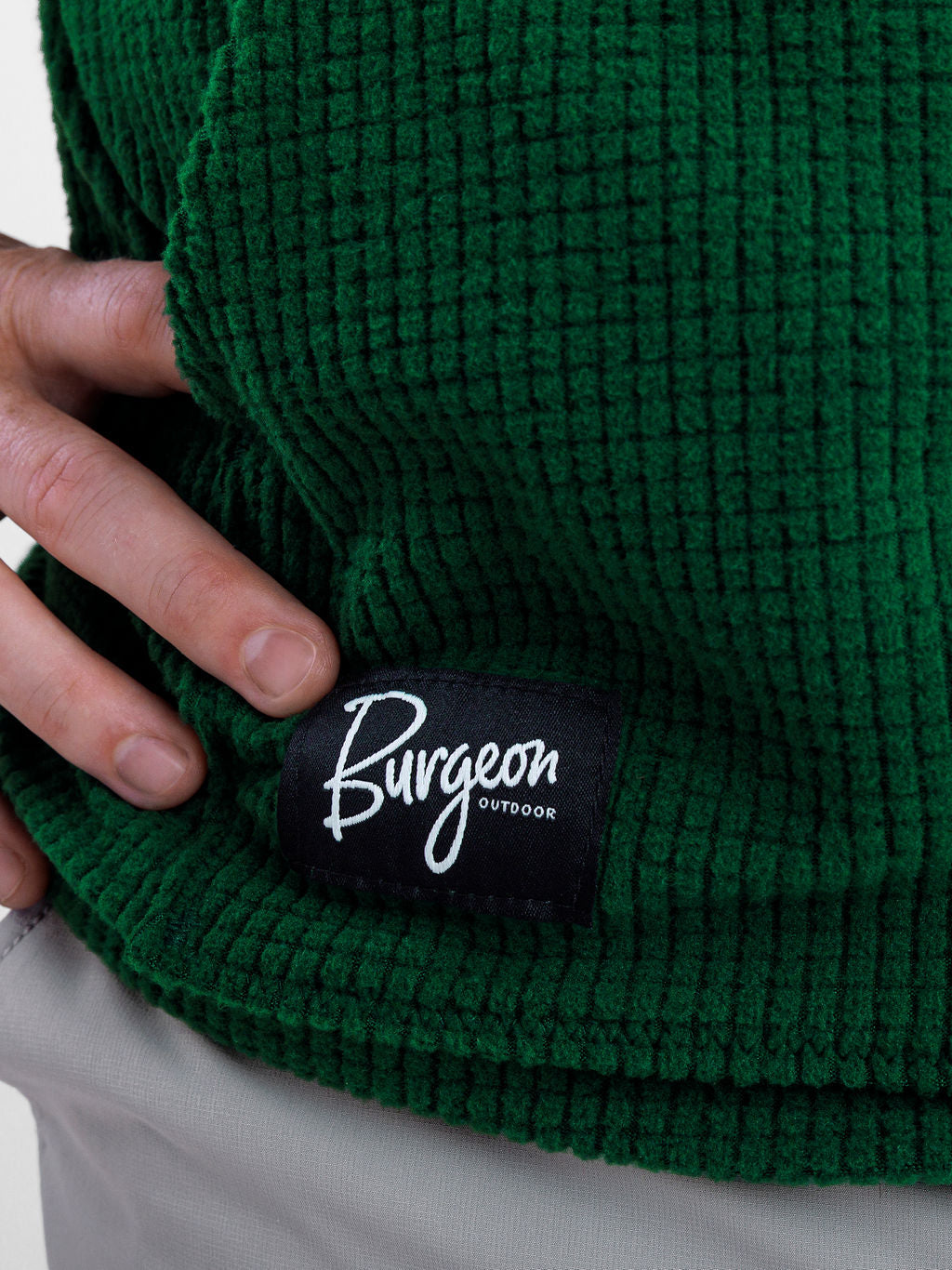 Close-up of a dark green Highlander fleece hoodie with a black sewn-on patch reading 'Burgeon OUTDOOR' in white script, worn by a person whose hand is touching the fabric.