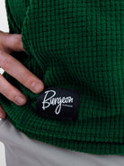 Close-up of a dark green Highlander fleece hoodie with a black sewn-on patch reading 'Burgeon OUTDOOR' in white script, worn by a person whose hand is touching the fabric.