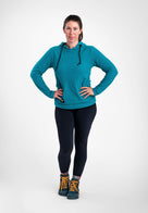 A woman modeling a teal Highlander hoodie with black drawstrings, paired with black leggings and hiking boots, standing against a light gray studio background.
